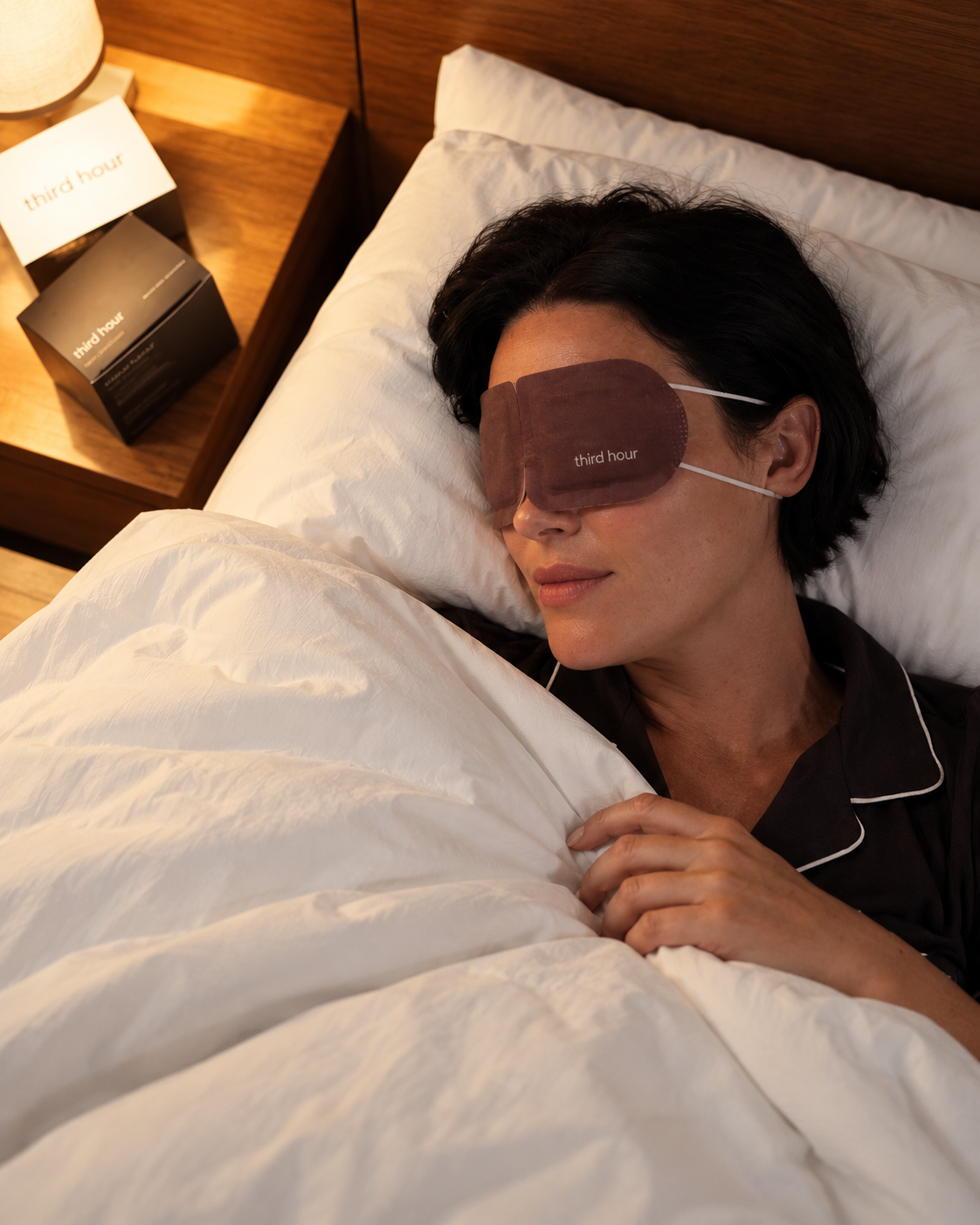 Woman resting in bed wearing the Third Hour warming sleep mask, with the Third Hour box on the bedside table